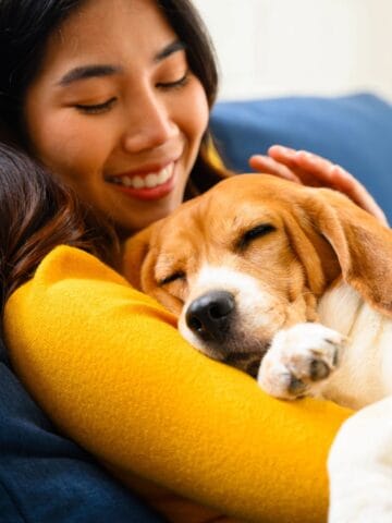 A relaxed woman smiles on the couch as her dog naps curled up against her arm.