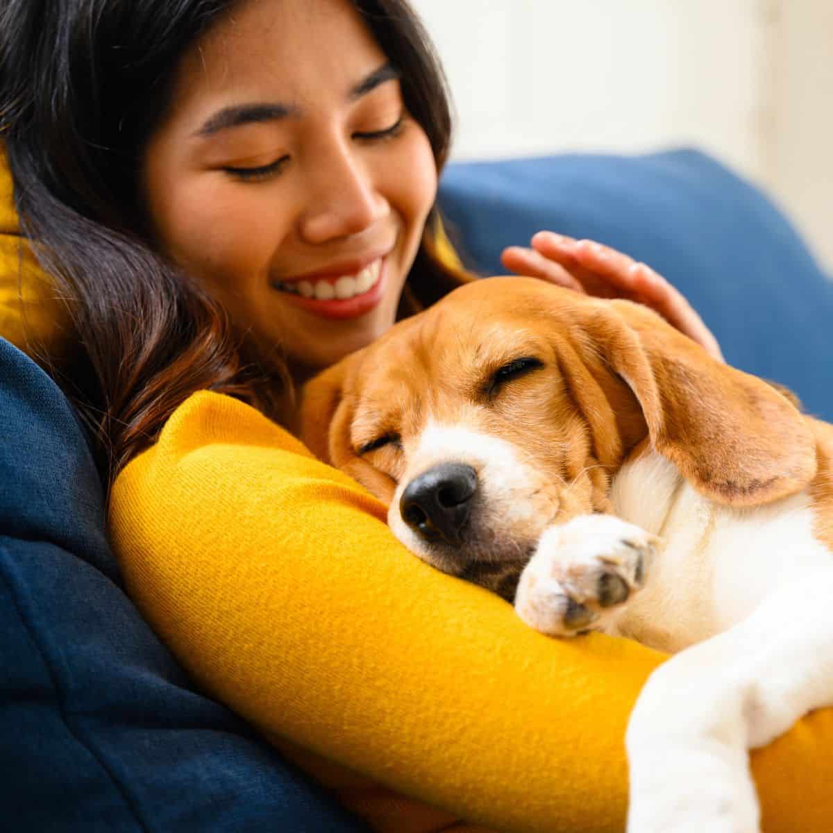 A relaxed woman smiles on the couch as her dog naps curled up against her arm.