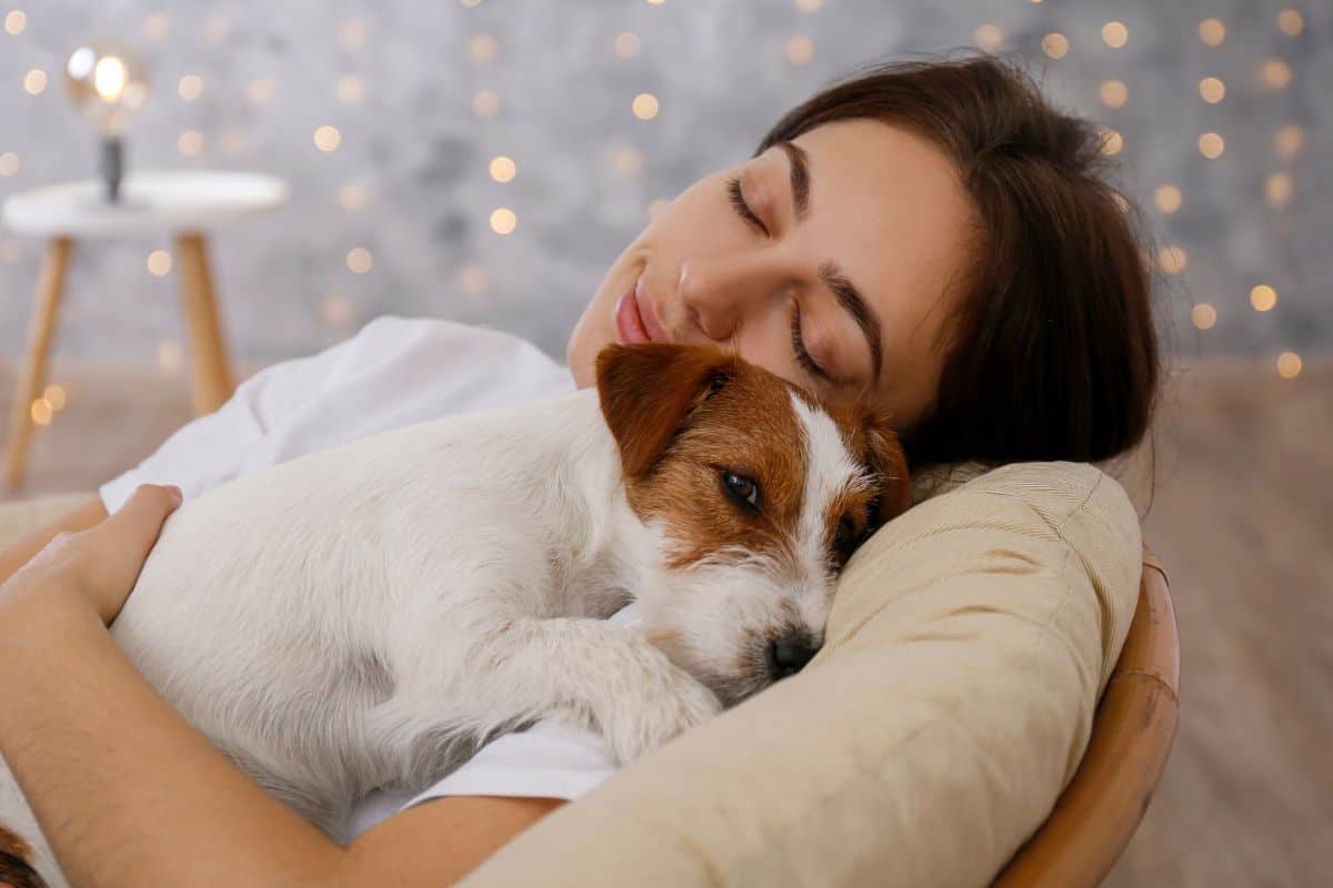 A woman smiling with her eyes closed while cuddling a small dog resting on her chest.