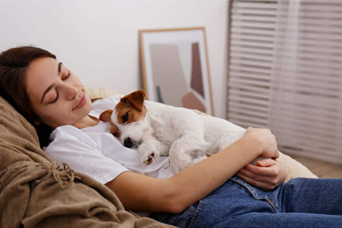 A woman relaxing on a couch while her small dog sleeps comfortably on her chest.