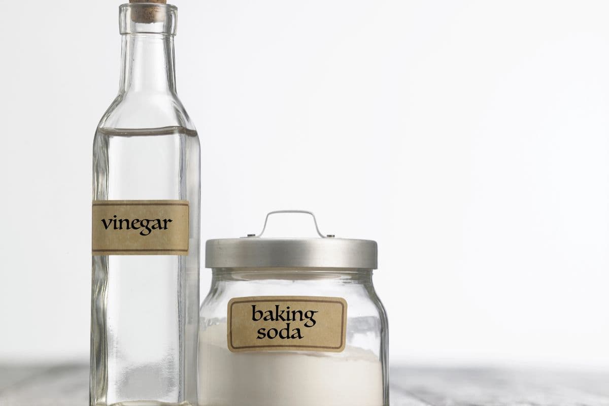 A glass bottle of vinegar and a jar of baking soda sit on a wooden surface against a white background.