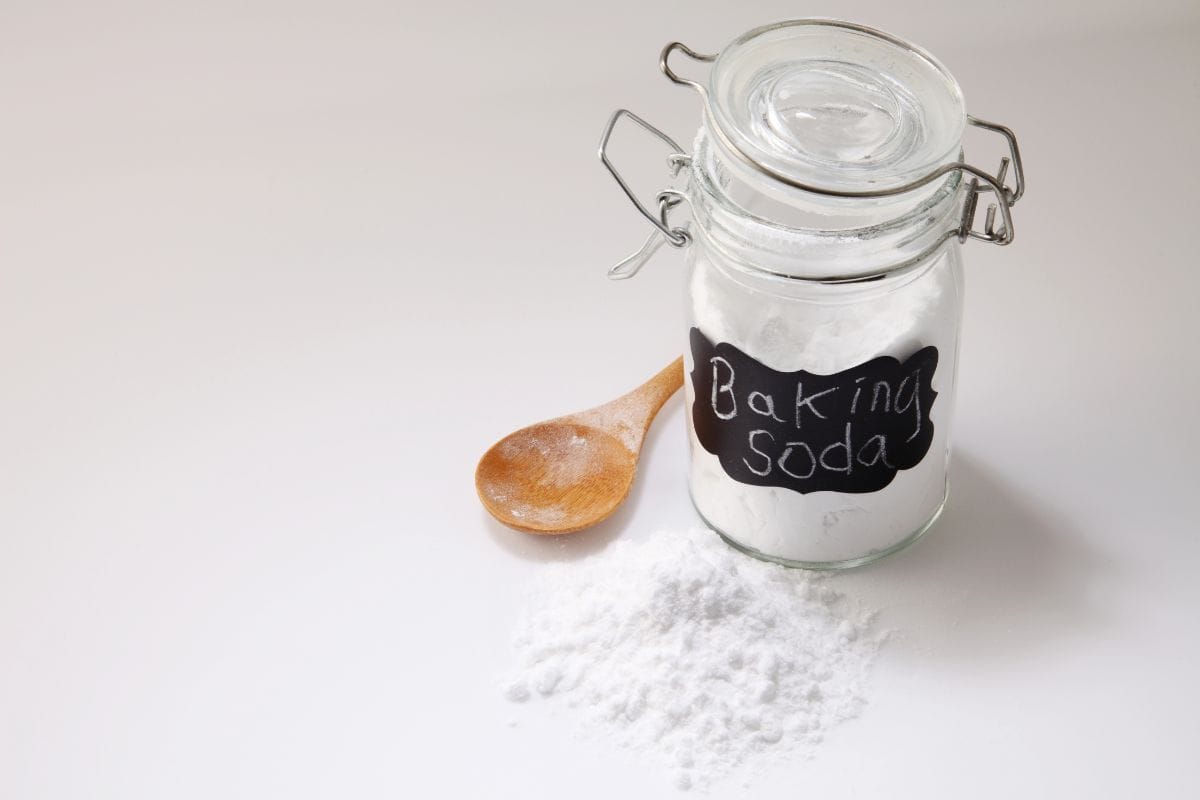 A glass jar labeled baking soda with a wooden spoon and spilled powder on a white surface.