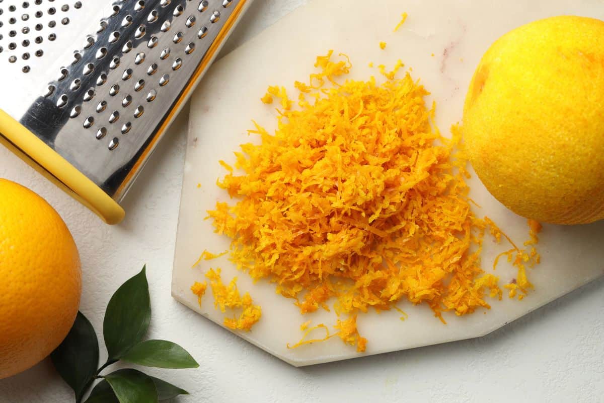Fresh orange zest piled on a cutting board beside whole oranges and a metal grater.