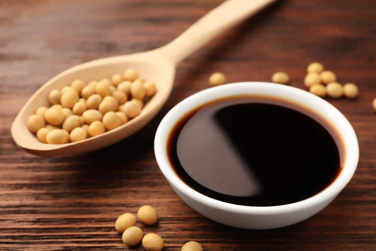 Bowl of soy sauce next to soybeans on a wooden surface.