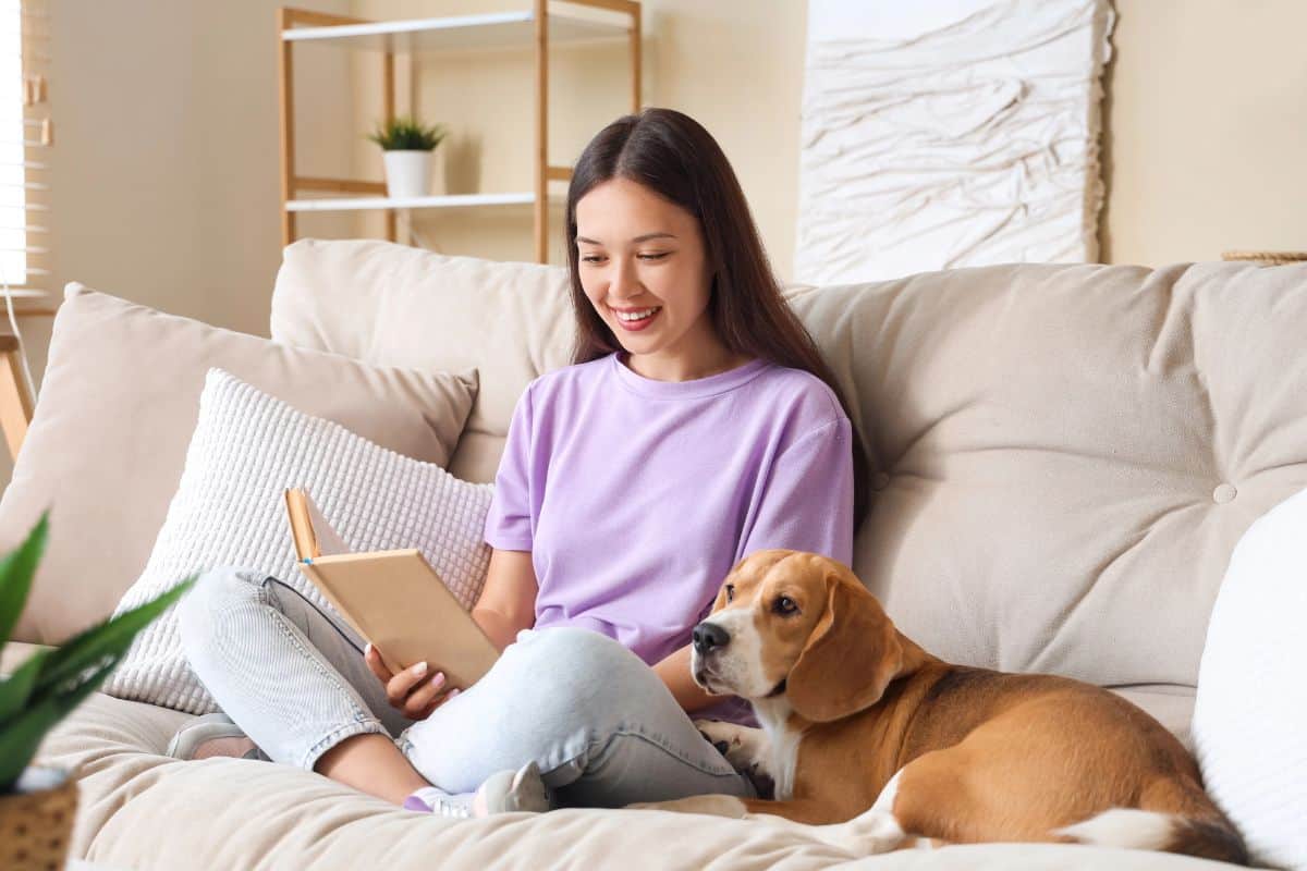 Woman reading a book on a couch with her dog beside her.