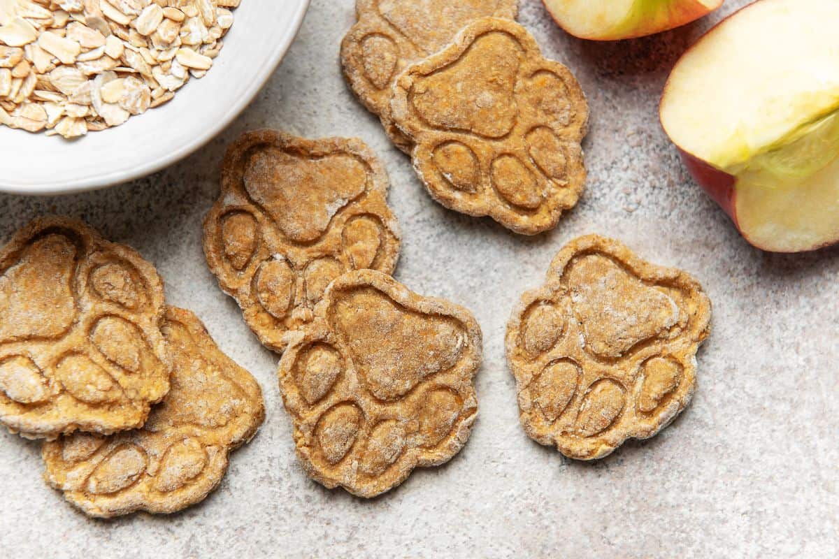 Paw-shaped gluten-free dog treats placed near oats and sliced apples.