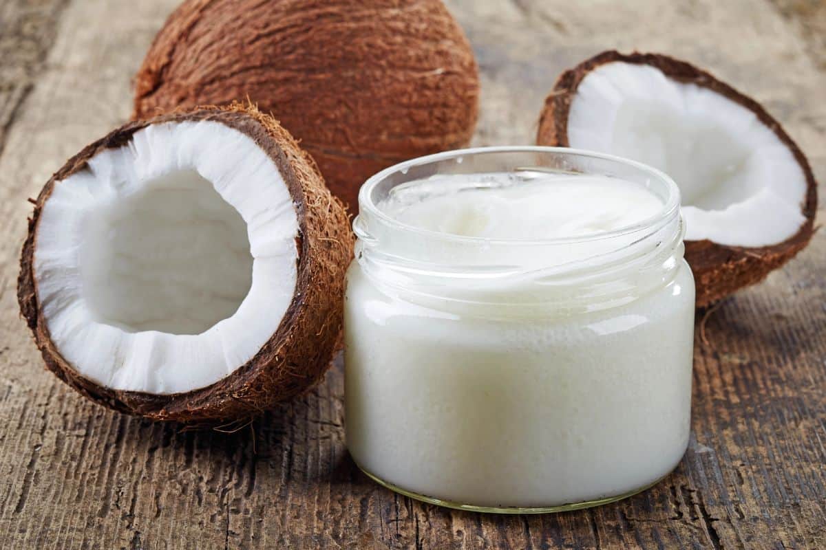 Jar of coconut oil beside fresh coconut halves on a wooden table.
