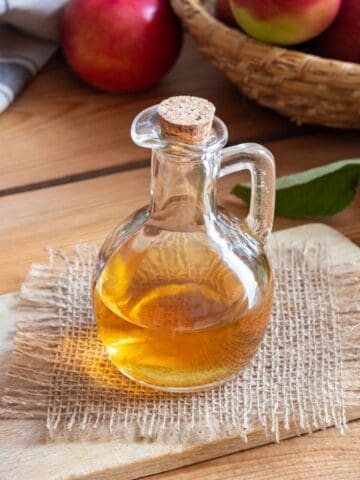 Glass bottle of apple cider vinegar surrounded by fresh apples on a wooden table.