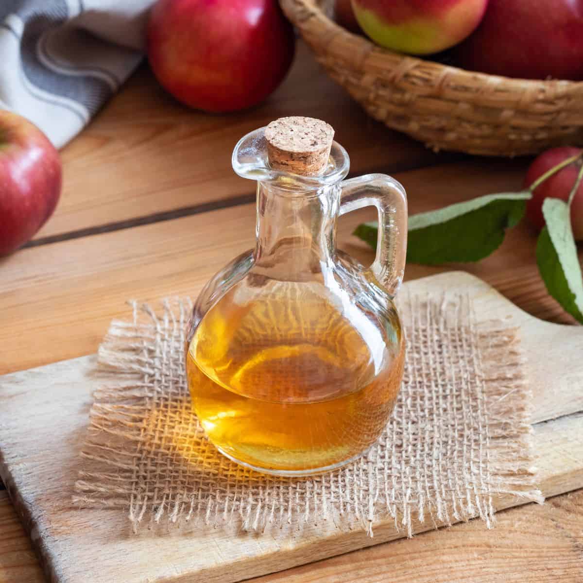 Glass bottle of apple cider vinegar surrounded by fresh apples on a wooden table.