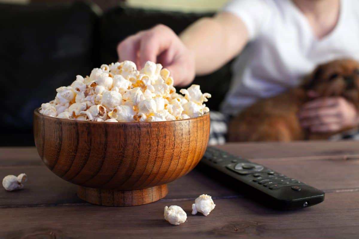 Person reaching for popcorn while holding a small dog on a couch.