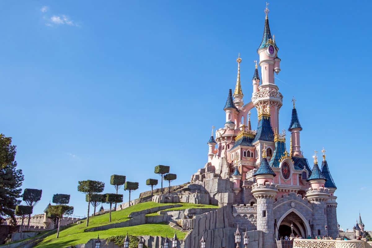 Disney castle with pink towers on a grassy hill under a clear blue sky.