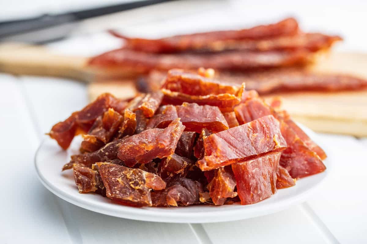Freshly cut dog jerky treats stacked on a white plate with whole jerky strips resting on a cutting board behind.