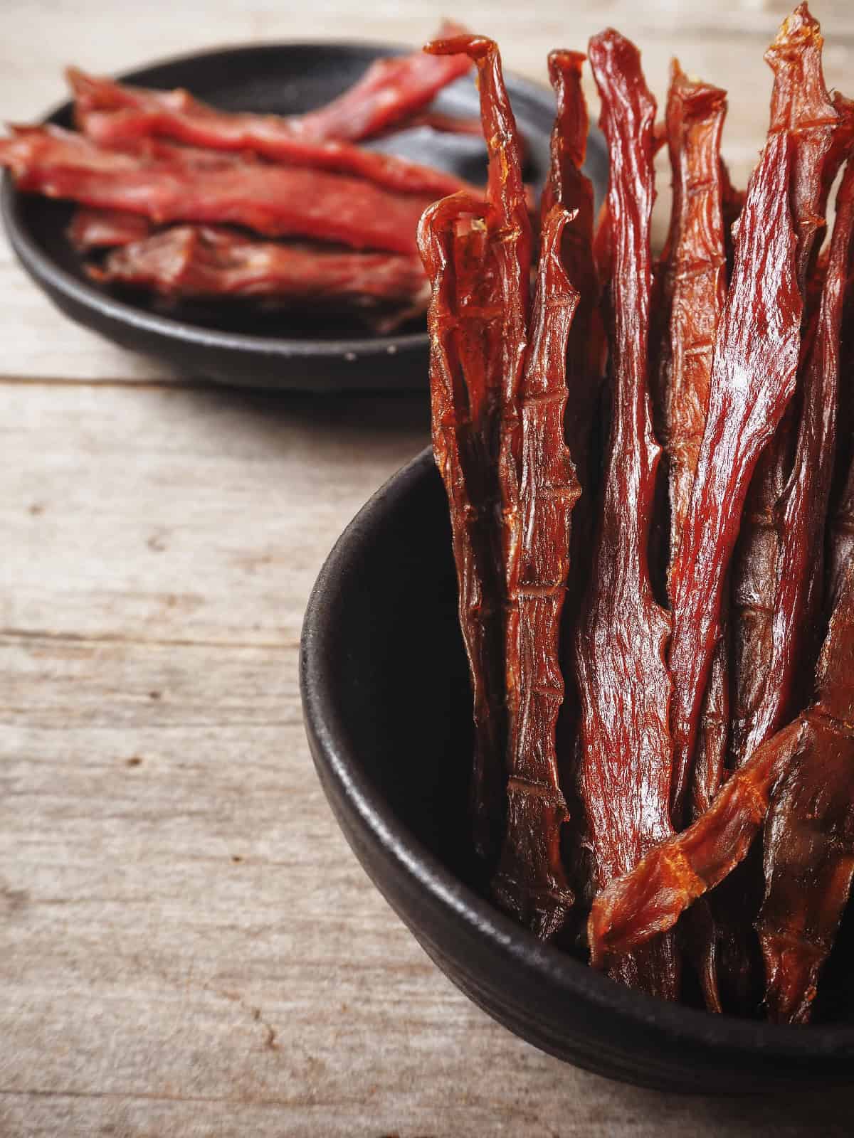 Long, chewy dog jerky strips standing upright in a black bowl with extra treats displayed on a plate in the background.