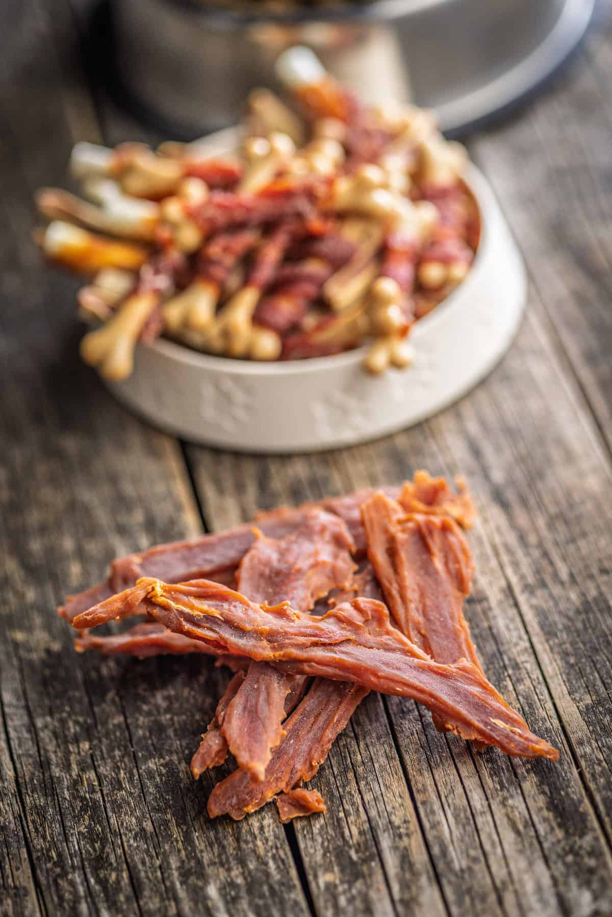 Homemade dog jerky strips on a rustic wooden table with a bowl of mixed dog treats blurred in the background.
